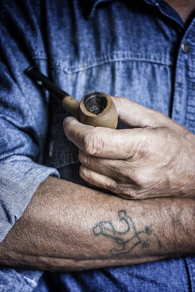 close up of an older man's folded arms where in one hand he holds a pipe and the other he shows a faded tattoo